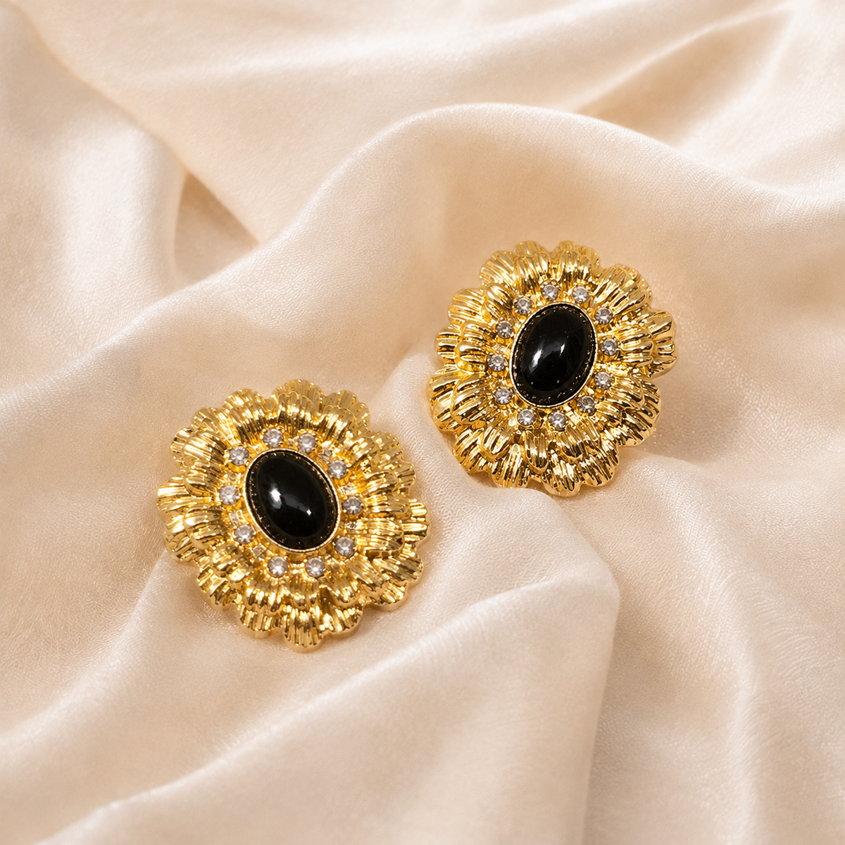 Pair of gold-tone radiating flower earrings with dark oval centers and clear round accents on warm cream silk